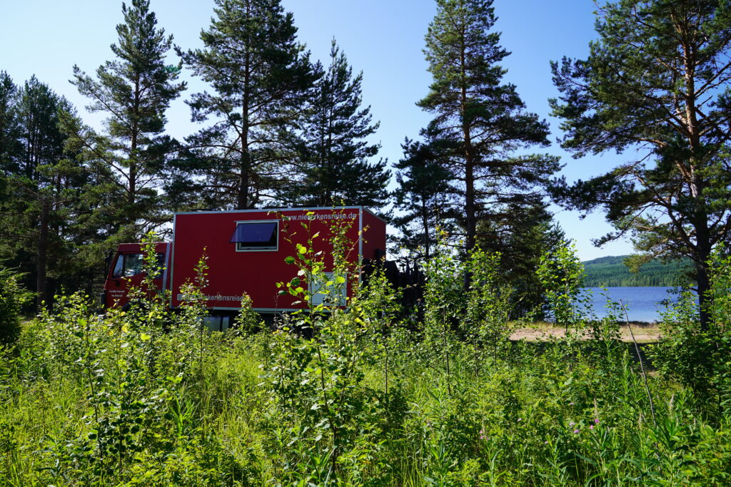 Roter Feuerwehr-LKW am Badeplatz Brintbordana in Schweden, umgeben von Natur, Blumen und Blick auf den See.