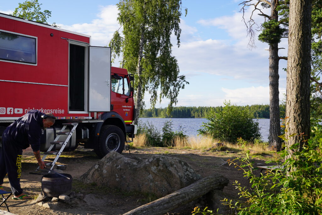 Feuerwehr-LKW am Ekna Naturcampingplatz direkt am See, zwischen Bäumen mit Feuerstelle im Vordergrund.