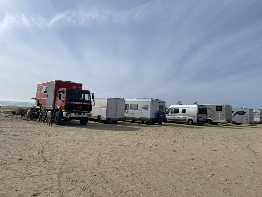 Umgebauter Feuerwehr LKW und mehrere Wohnmobile auf einem Parkplatz direkt am Strand vor Cadiz, Spanien