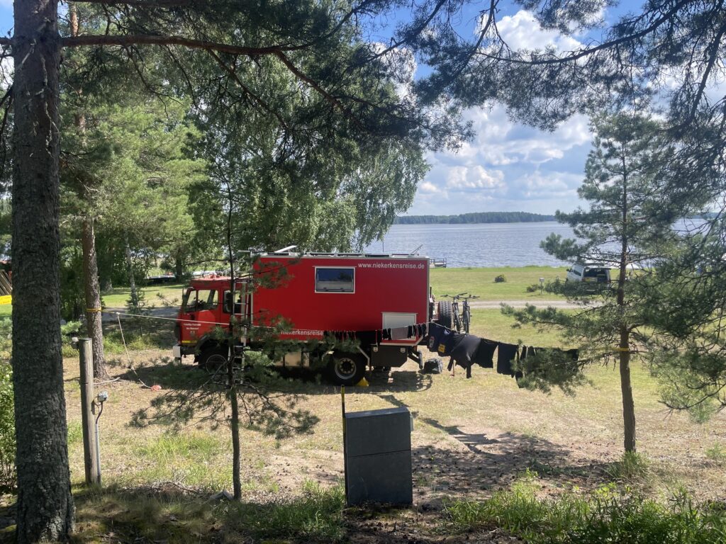 Roter Feuerwehr-LKW als Camper am Campingplatz Schwedenpause mit Wäschleine unter Bäumen direkt am See.“