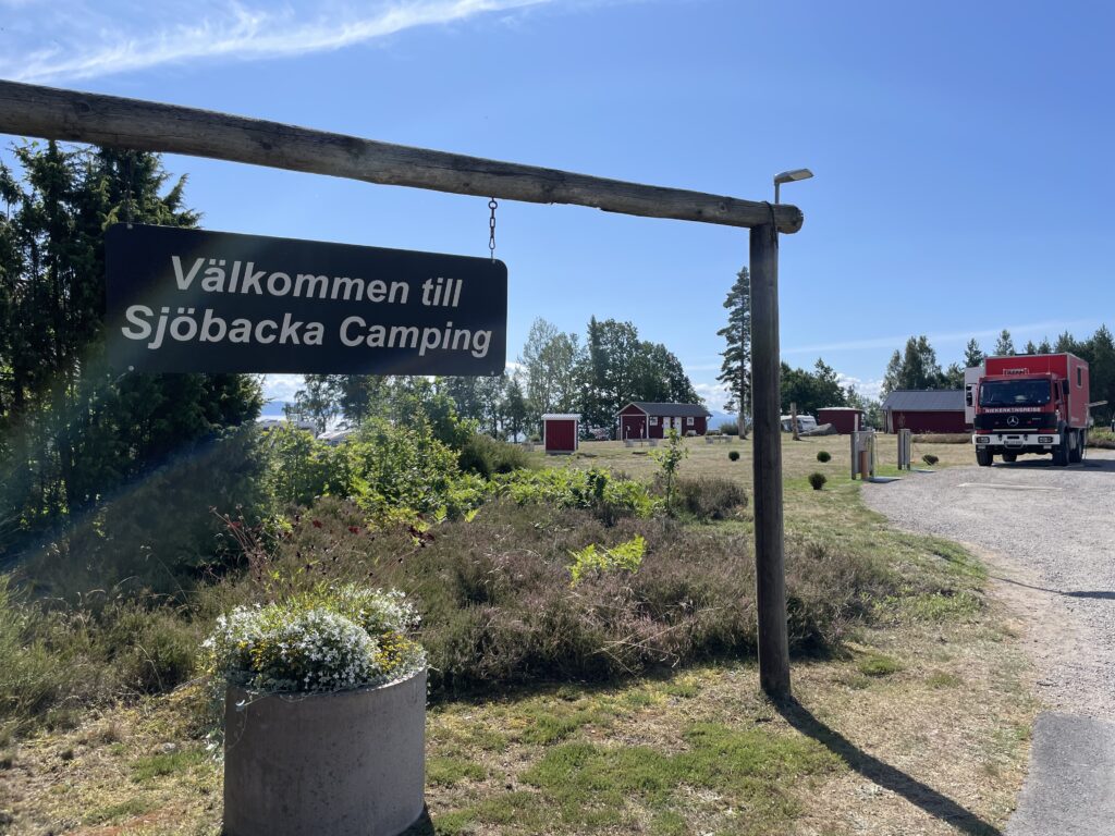 Einfahrt zum Sjöbacka Camping in Schweden mit Willkommensschild und rotem Feuerwehr-LKW auf dem Platz.