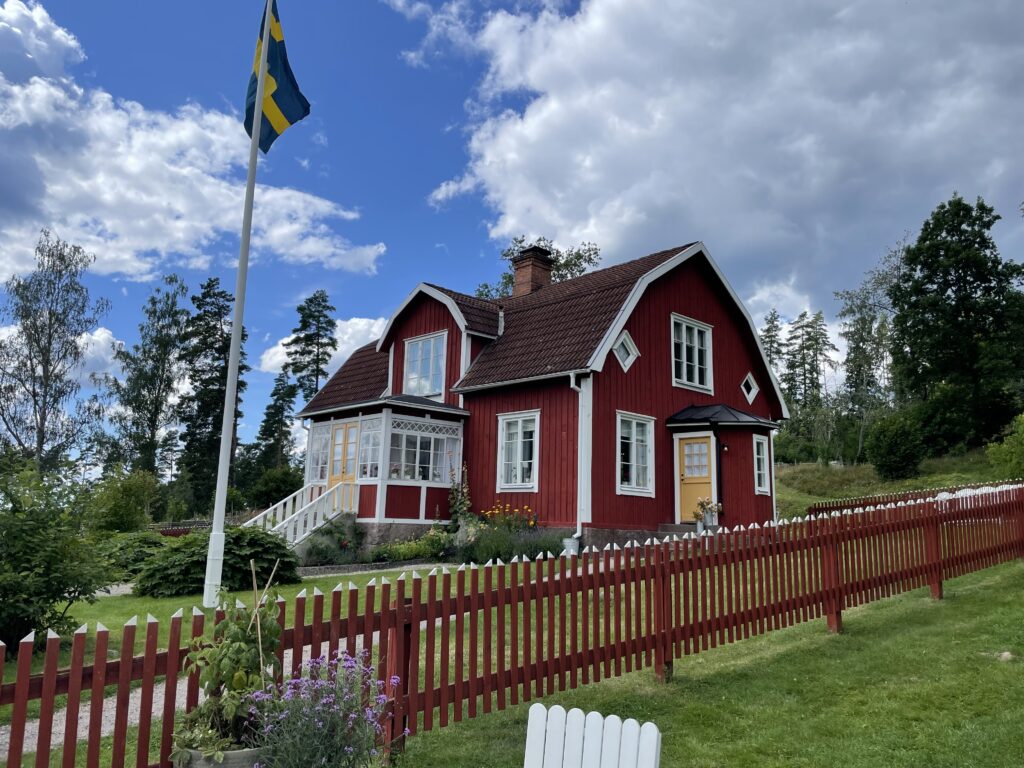 Rotes Schwedenhaus, der berühmte Katthulthof aus Michel aus Lönneberga mit schwedischer Flagge, Garten und weißem Zaun bei blauem Himmel.