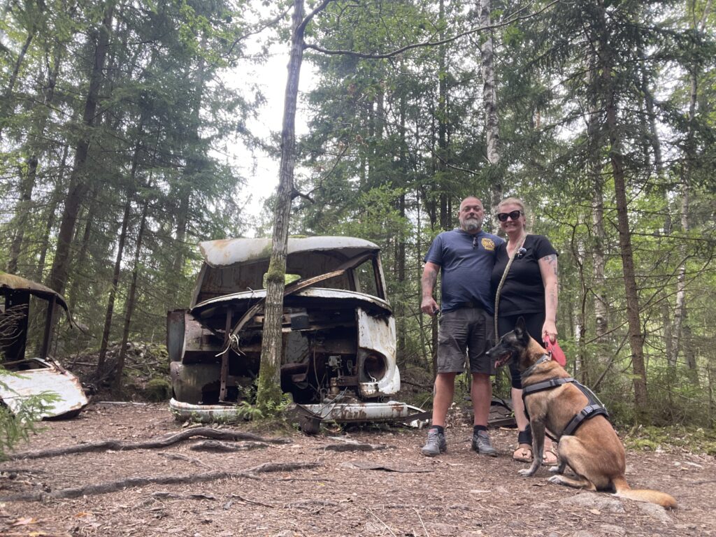 Zwei Erwachsene mit Hund vor einem verrosteten Autowrack im Wald am Autofriedhof Kyrkö Mosse in Schweden.