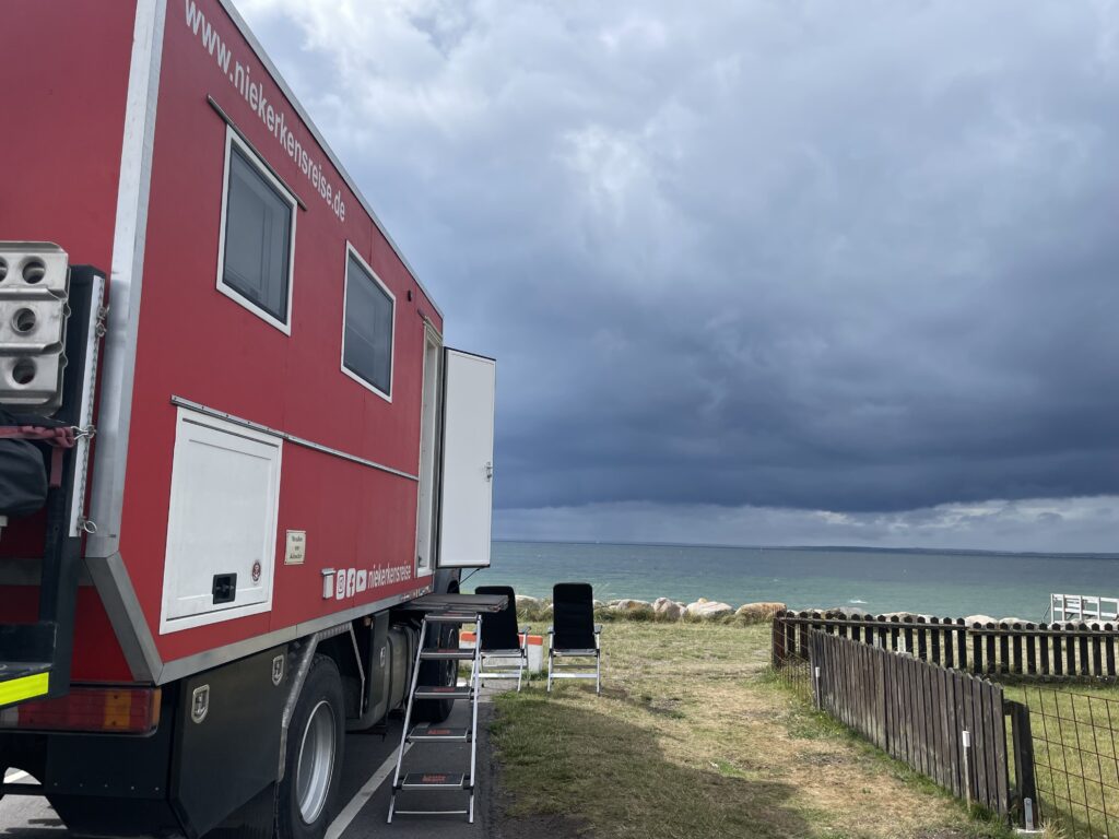 Roter Feuerwehr-LKW als Wohnmobil am Stellplatz im Sportboothafen Helsingborg mit Blick aufs Meer unter dunklen Wolken.