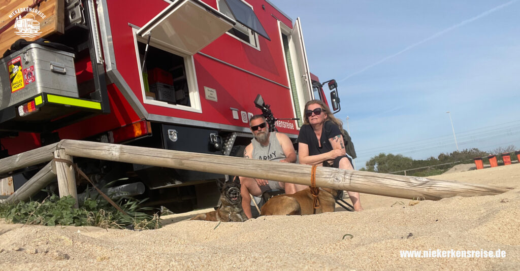 Anja und Lars von niekerkensreise sitzen mit ihren Hunden vor ihrem selbstausgebauten Feuerwehr-LKW Mercedes Benz 1224 an einem Stellplatz in Strandnähe
