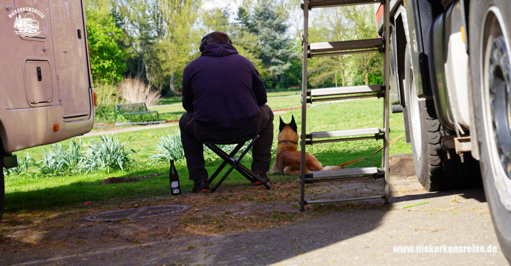 Lars sitzt mit Hund vor dem Expeditionsmobil von niekerkensreise auf dem Reisemobilhafen Köln und genießt die ruhige Atmosphäre im Grünen