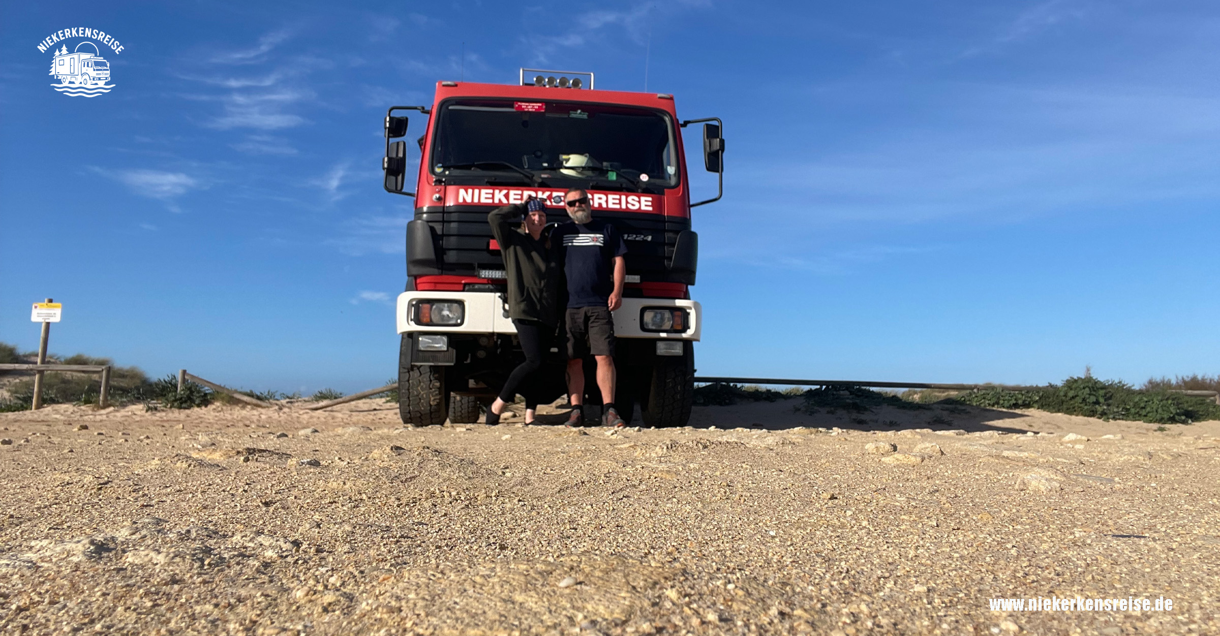 Freier Stellplatz bei Cádiz mit rotem Expeditionsmobil und zwei Personen - Anja und Lars von niekerkensreise - vor dem Fahrzeug an der Küste
