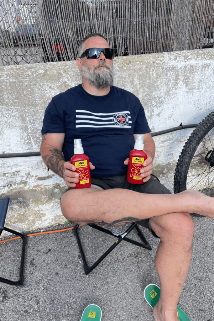 Lars sitzt entspannt auf einem Campingstuhl und hält zwei Flaschen Anti Brumm Forte Mückenspray in der Hand – effektiver Mückenschutz beim Camping und Vanlife.