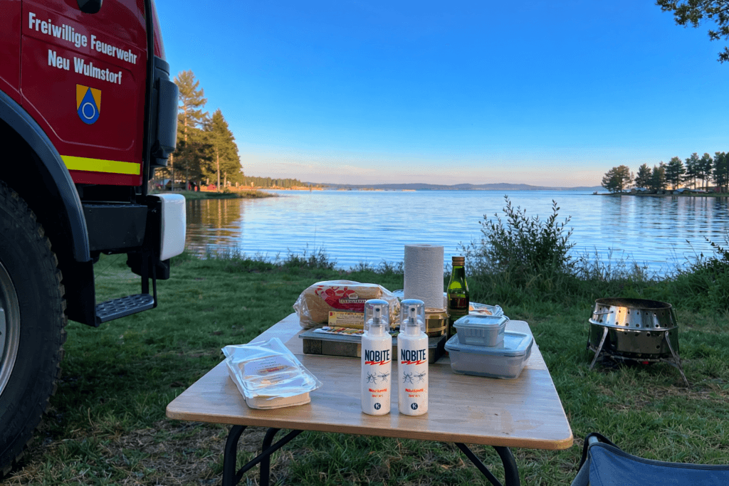 Nobite Mückenspray auf Campingtisch am See neben Expeditionsmobil – effektiver Mückenschutz beim Camping und Vanlife in der Natur.