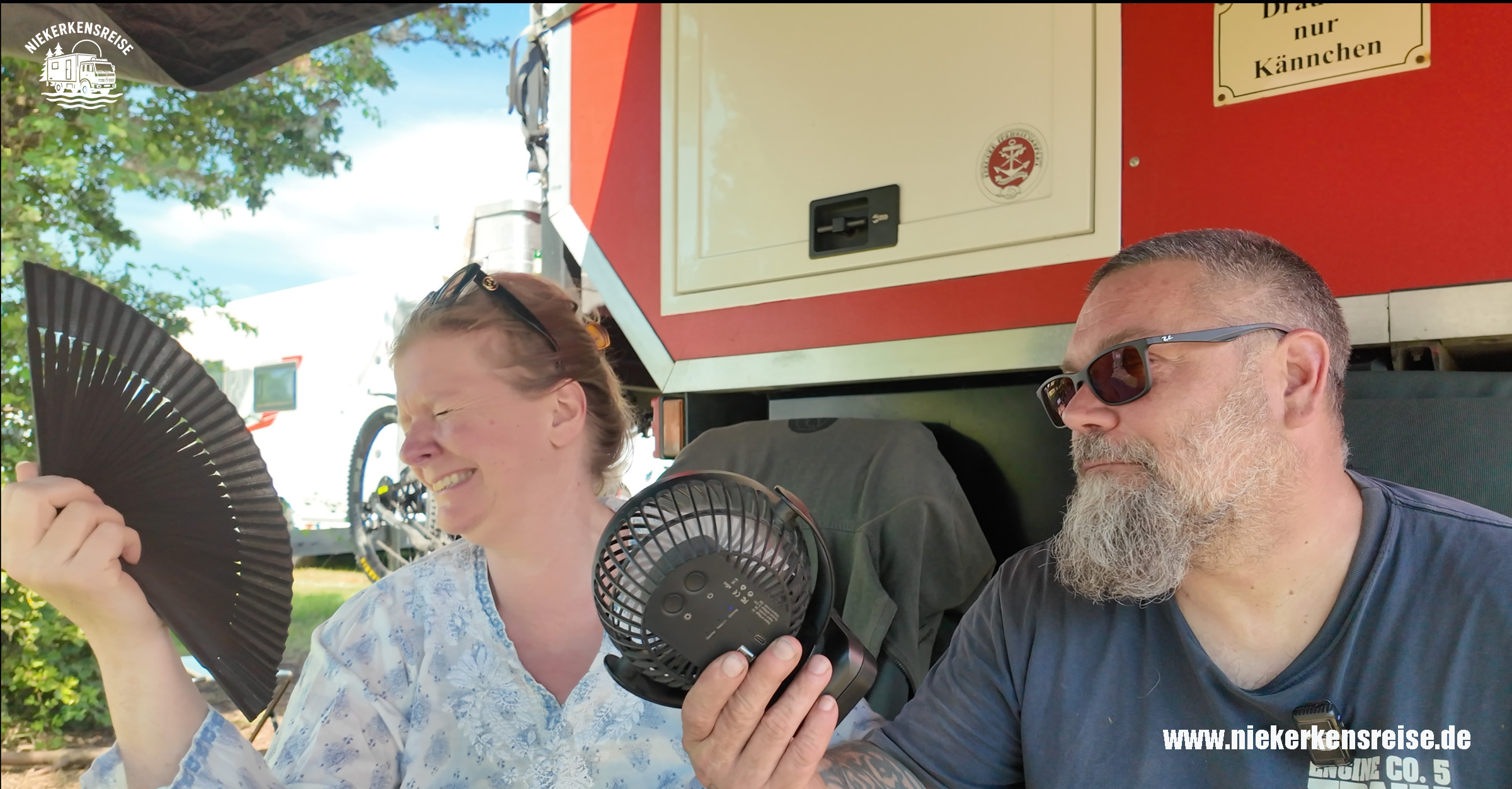 Anja und Lars kühlen sich mit Handfächer und USB-Ventilator vor ihrem Expeditions-Wohnmobil an einem heißen Sommertag ohne Klimaanlage.
