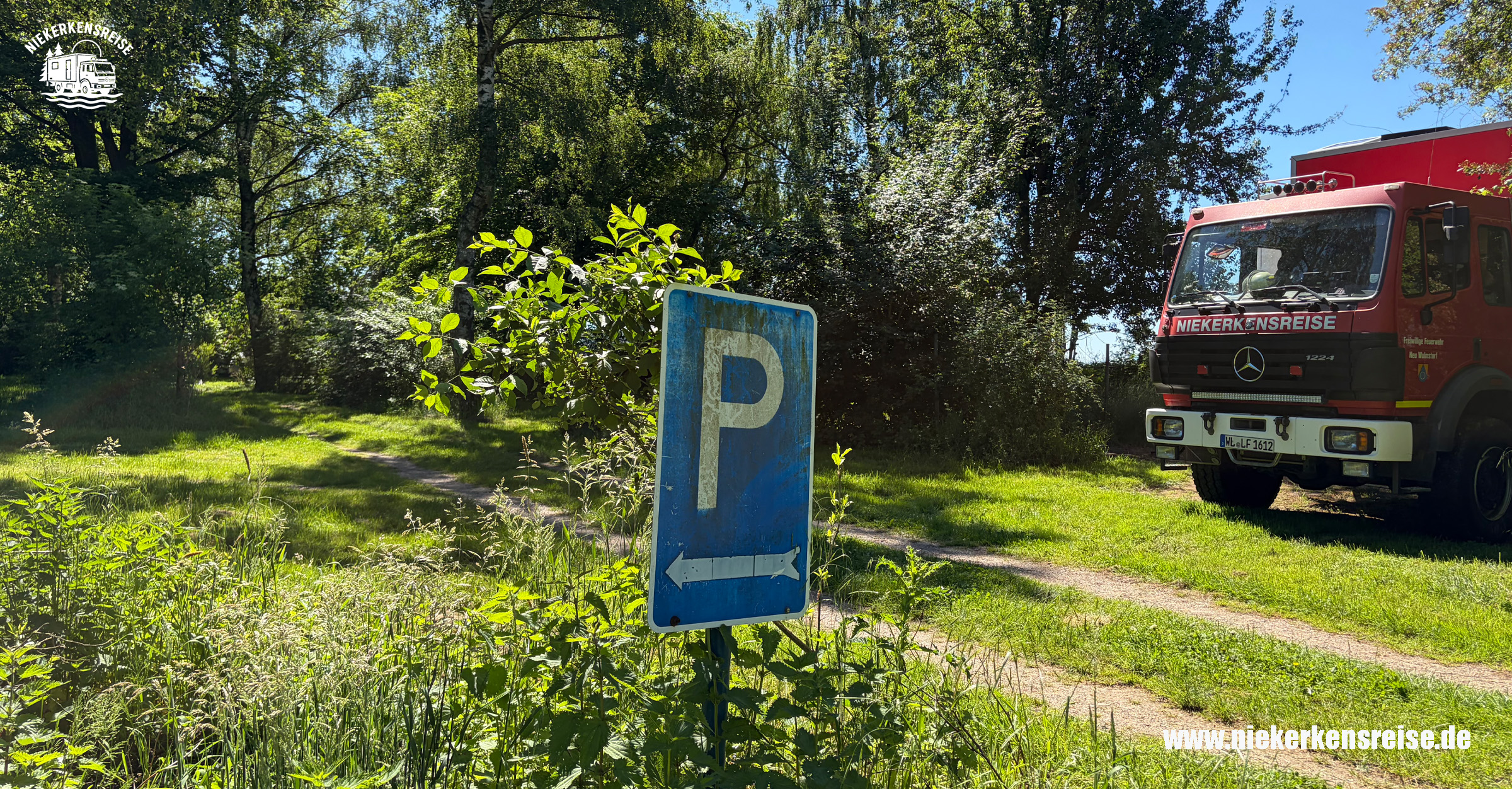 Selbst ausgebauter Feuerwehr-LKW von Niekerkensreise auf einem schattigen Naturstellplatz an der Weser. Das Expeditionsmobil steht zwischen großen Bäumen auf einer grünen Wiese neben einem Parkplatzschild.