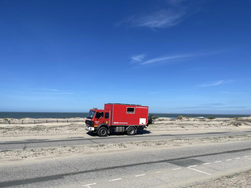 Rotes Expeditionswohnmobil auf einer Küstenstraße in Frankreich. Im Hintergrund liegen Sanddünen und das Meer unter einem strahlend blauen Himmel. Das Bild zeigt einen Roadtrip mit dem Wohnmobil entlang der Atlantikküste.