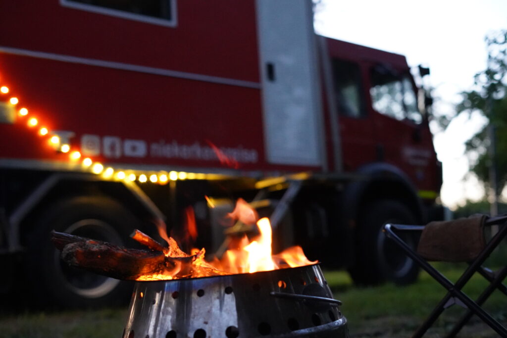 Feuerschale vor dem roten Expeditionsmobil von Niekerkensreise in der Abenddämmerung. Das knisternde Lagerfeuer und die dezente Außenbeleuchtung schaffen eine gemütliche Campingatmosphäre auf dem Stellplatz.
