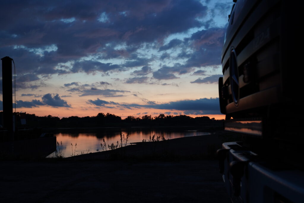 Mercedes-Benz Expeditionsmobil von Niekerkensreise am Ufer eines ruhigen Flusses in der Abenddämmerung. Die friedliche Stimmung bei Sonnenuntergang symbolisiert die Bedeutung von Ruhezeiten auf dem Campingplatz.