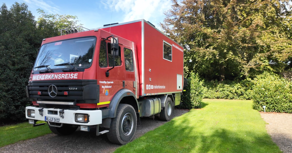 Roter Mercedes-Benz 1224 AF Expeditions-Lkw von Niekerkensreise auf einer geschotterten, von Rasen und Hecken umgebenen Stellfläche in Kessel-Eik, Niederlande.
