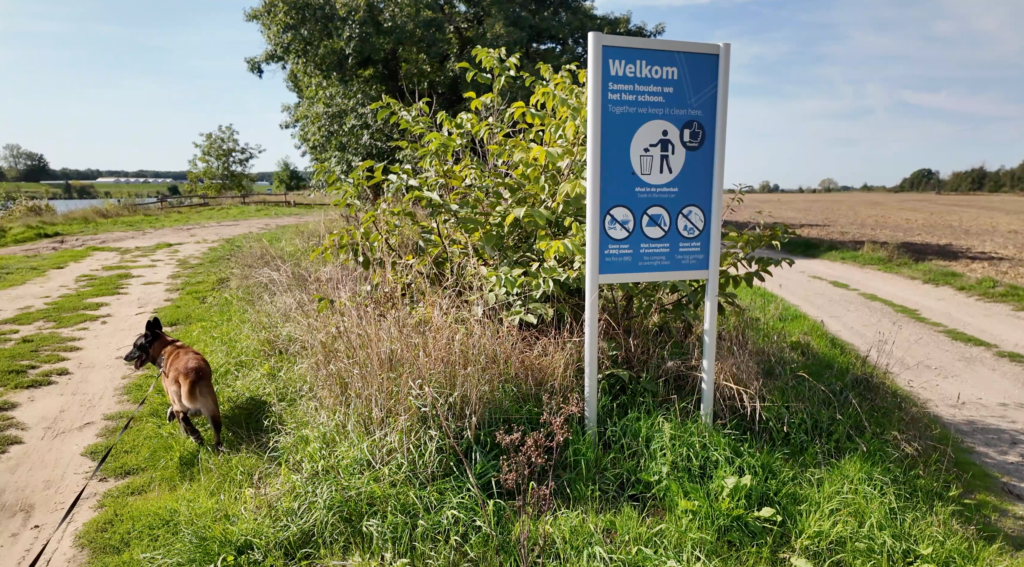 Malinois Elvis beim Spaziergang an der Maas bei Kessel-Eik in Limburg, neben einem Hinweisschild mit Verhaltensregeln für Besucher.