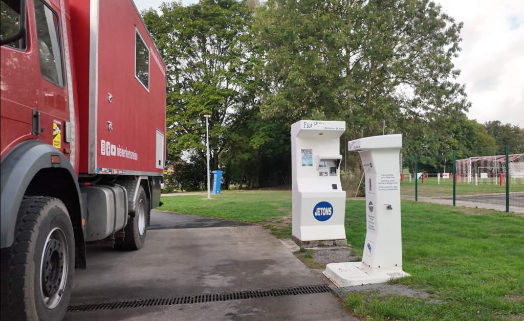Ver- und Entsorgungsstation auf dem kostenlosen Wohnmobilstellplatz Solre-le-Château in Nordfrankreich mit Frischwassersäule, Jeton-Automat und rotem Expeditionsmobil von Niekerkensreise.