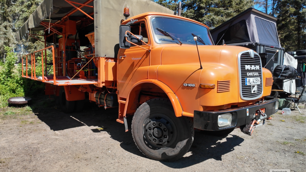 Orangefarbener MAN 13.168 Diesel Oldtimer-LKW mit historischem Arbeitsaufbau und seitlicher Plattform beim Overlander-Treffen Quaduxenbarg 2026.