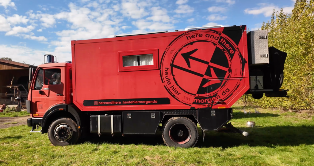 Roter Mercedes-Benz 1222 „Otto“ von Katrin und Andre mit selbst gebauter Wohnkabine aus Holz, Allradantrieb und großem „Here and There“-Logo auf der Fahrzeugseite.