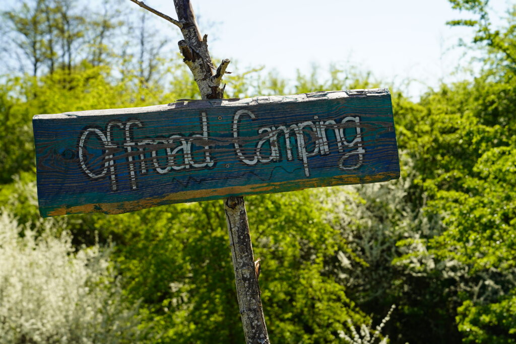 Rustikales, blau-grünes Holzschild mit der Aufschrift „Offroad Camping“ an einem Baum auf dem Quaduxenbarg, umgeben von grüner Natur beim Overlander-Treffen 2026.