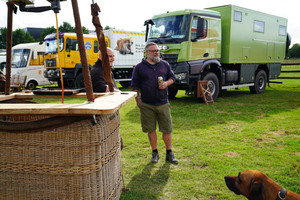 Zwei Teilnehmer beim Hof & Hubraum Treffen 2026 vor einem grünen Mercedes-Benz Arocs Expeditionsmobil. Im Vordergrund steht ein großer Korbgrill, rechts ist ein Hund zu sehen. Im Hintergrund parken weitere Wohnmobile und Allrad-LKW.