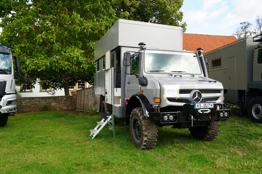 Grauer Mercedes-Benz Unimog mit hoher Hubdach-Wohnkabine und grobstolligen Geländereifen beim Hof & Hubraum Treffen 2026. Der individuell ausgebaute Expeditions-LKW steht auf einer Wiese.