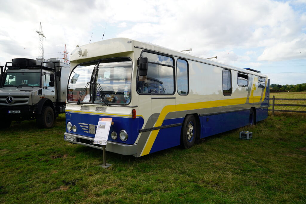 Historischer Büssing Bus von 1962 mit blau-weiß-gelber Lackierung, zum Wohnmobil selbst ausgebaut, beim Hof & Hubraum Treffen 2026. Der Oldtimer steht auf einer Wiese neben einem Expeditions-LKW.