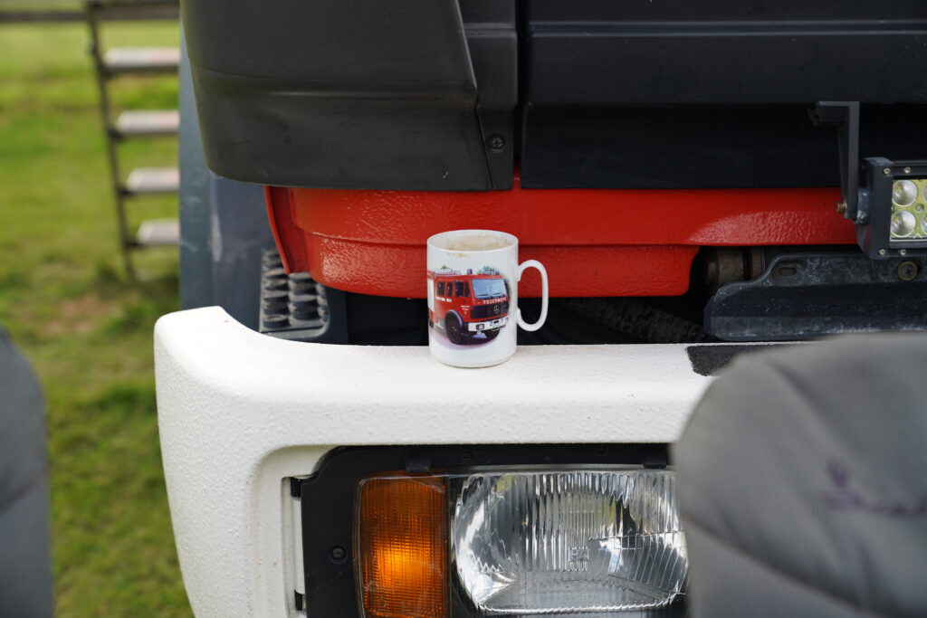 Weiße Kaffeetasse mit dem Motiv eines roten Mercedes-Benz Feuerwehr-LKW auf der Stoßstange eines Expeditionsmobils beim Hof & Hubraum Treffen 2026.