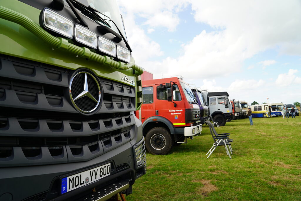 Grüner Mercedes-Benz Arocs Allrad-LKW im Vordergrund und mehrere Expeditionsmobile, darunter ehemalige Feuerwehrfahrzeuge und ein historischer Bus, beim Hof & Hubraum Treffen 2026 auf einer Wiese.