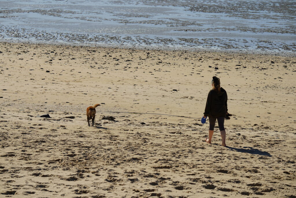 Anja spaziert barfuß mit Hund Elvis am weitläufigen Sandstrand von Dragey-Ronthon in der Normandie. Bei Ebbe erstreckt sich die Bucht des Mont-Saint-Michel mit Sandflächen und Watt bis zum Horizont.