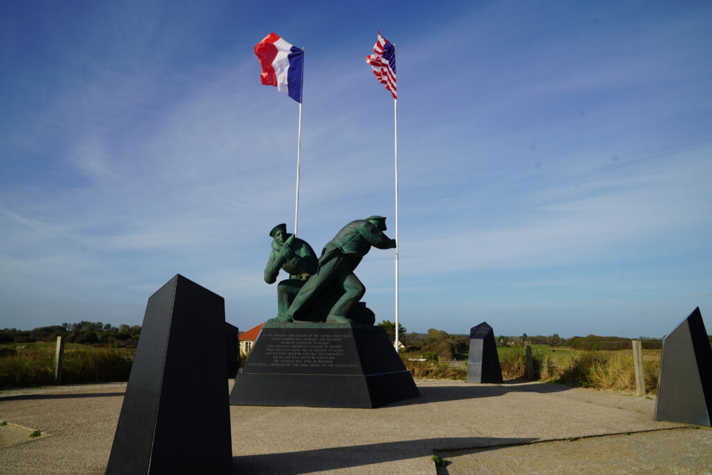 Denkmal der US-Marine am Utah Beach in der Normandie mit Bronzeskulpturen amerikanischer Marinesoldaten. Im Hintergrund wehen die französische und die amerikanische Flagge am historischen Landungsstrand des D-Day.