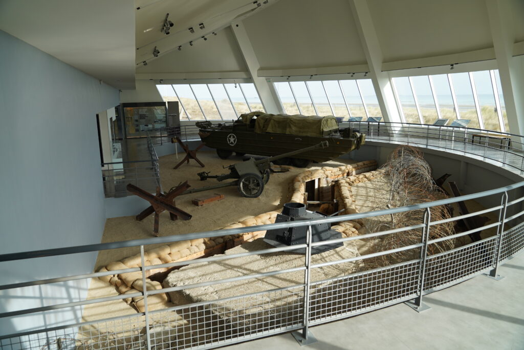 Ausstellung im Utah Beach Landing Museum in der Normandie mit historischem US-Militärfahrzeug, Panzerabwehrkanone, Sandsäcken und Stacheldraht. Durch die großen Panoramafenster ist der ehemalige Landungsstrand des D-Day zu sehen.