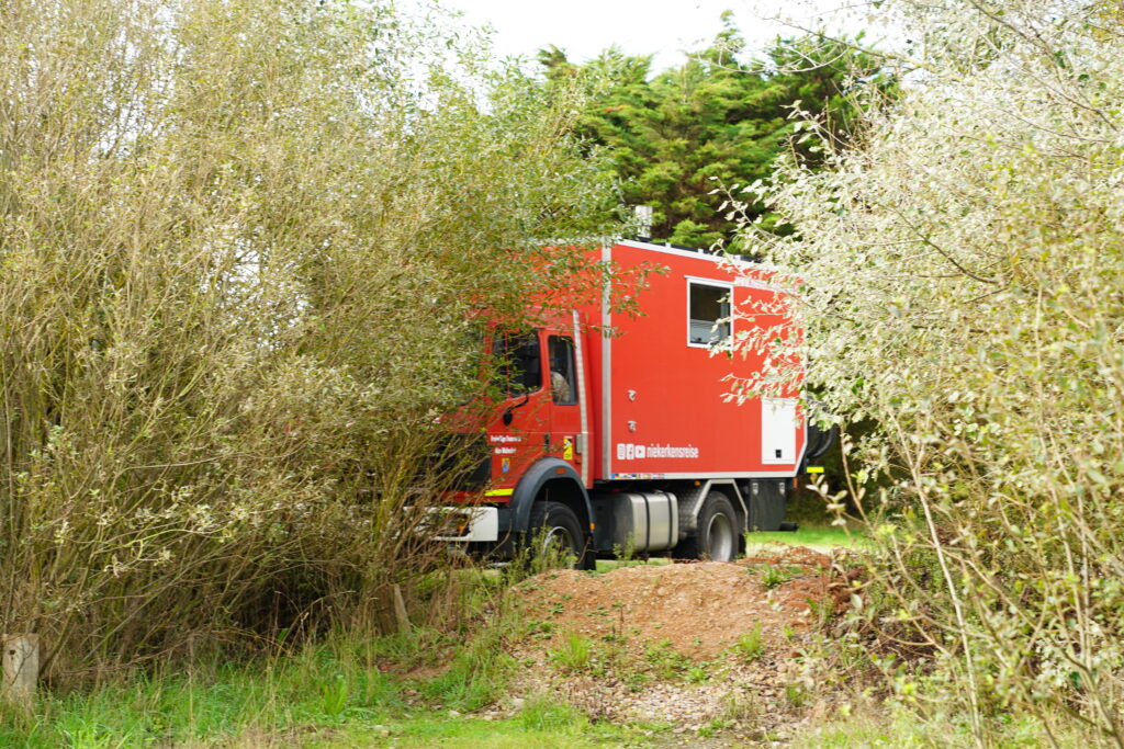 Roter Mercedes-Benz Expeditions-LKW von Niekerkensreise auf dem kostenlosen Wohnmobilstellplatz am Utah Beach in der Normandie. Das Wohnmobil steht auf einer von Bäumen und Sträuchern umgebenen Stellfläche im Grünen.