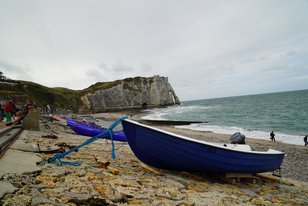 Blaue Fischerboote am Kieselstrand von Étretat in der Normandie. Im Hintergrund erhebt sich die berühmte weiße Kreideklippe Falaise d’Aval mit ihrem natürlichen Felsbogen Porte d’Aval über dem Ärmelkanal.