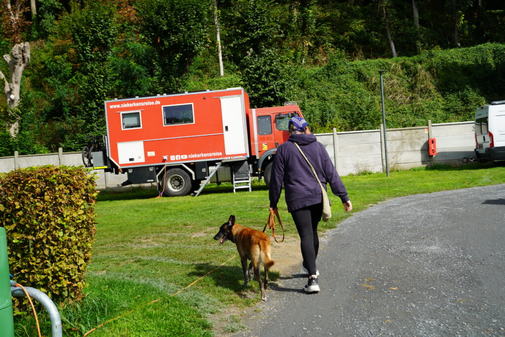 Anja spaziert mit Malinois Elvis über den Camping Municipal d’Étretat in der Normandie. Im Hintergrund steht der rote, selbst ausgebaute Mercedes-Benz Feuerwehr-LKW von Niekerkensreise auf einer grünen Stellplatzwiese.