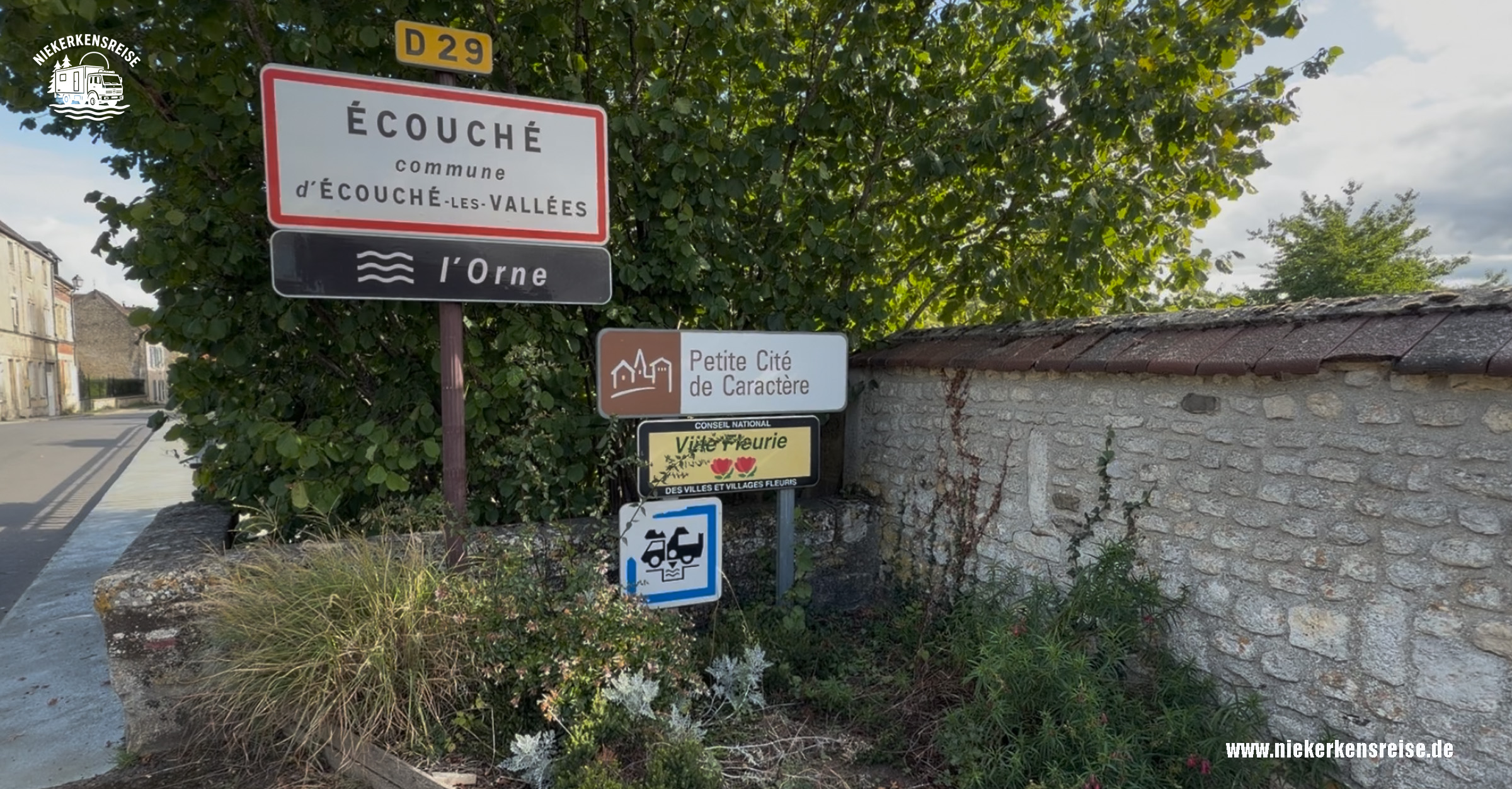 Écouché-les-Vallées – Historischer Ort mit Wohnmobilstellplatz in der Normandie Ortseingangsschild von Écouché-les-Vallées in der Normandie, Frankreich, mit Hinweisschildern auf die Petite Cité de Caractère und eine Wohnmobil-Servicestation.