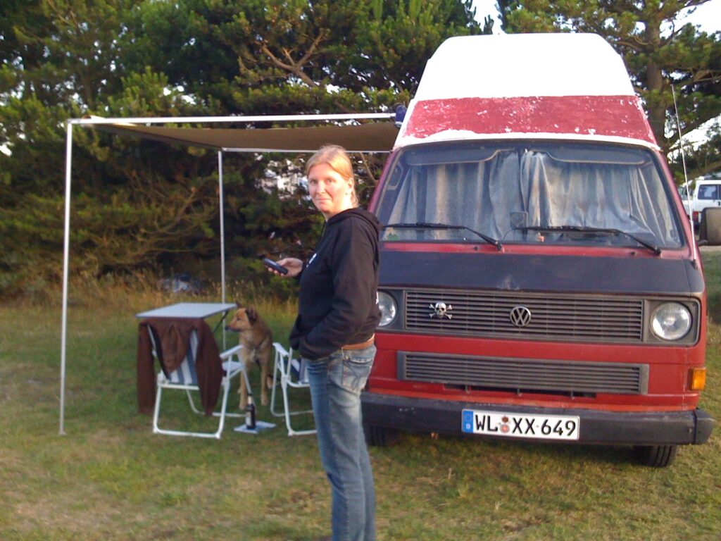 Anja steht vor dem ersten VW T3 Bulli von Niekerkensreise, einem rot-weißen Camper mit Weinsberg-Ausbau. Neben dem Fahrzeug sind ein Vorzelt, Campingstühle und ein Hund zu sehen.