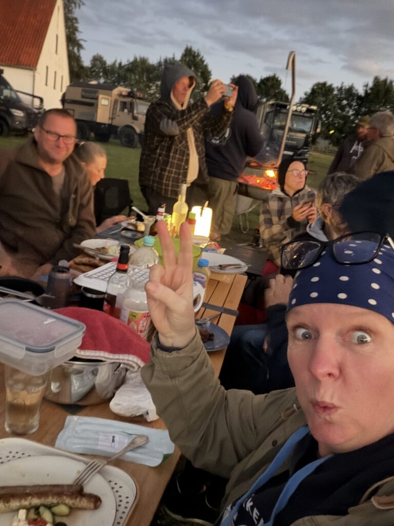 Gemütliches Beisammensein beim Hof & Hubraum Treffen 2026: Camper und Expeditionsmobil-Fahrer sitzen abends gemeinsam an einer langen Tafel, essen und unterhalten sich. Im Hintergrund stehen Allrad-LKW und ein Grill.