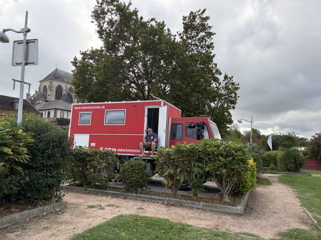 Rotes Mercedes-Benz 1224 AF Expeditionsmobil von Niekerkensreise auf dem Wohnmobilstellplatz in Pont-de-l’Arche in der Normandie. Im Hintergrund ist die historische Kirche Notre-Dame-des-Arts zu sehen.