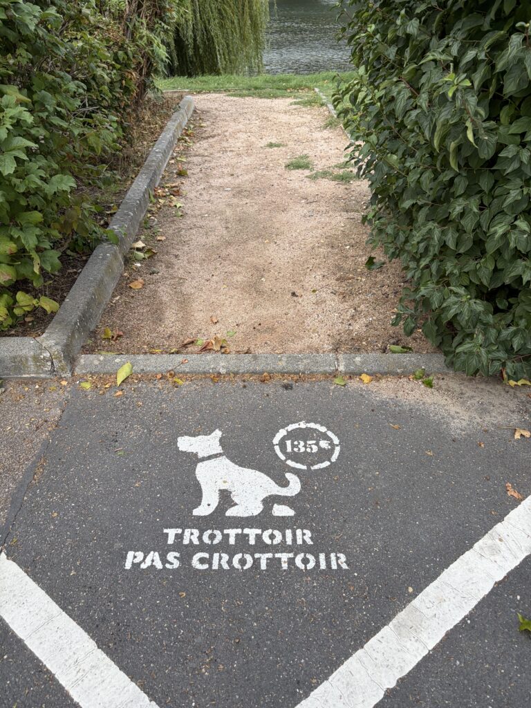 Hundefreundlicher Wohnmobilstellplatz in Pont-de-l’Arche in der Normandie. Eine französische Bodenmarkierung mit Hundesymbol und der Aufschrift „Trottoir pas crottoir“ weist auf die Sauberkeitspflicht am Fußweg zur Eure hin.