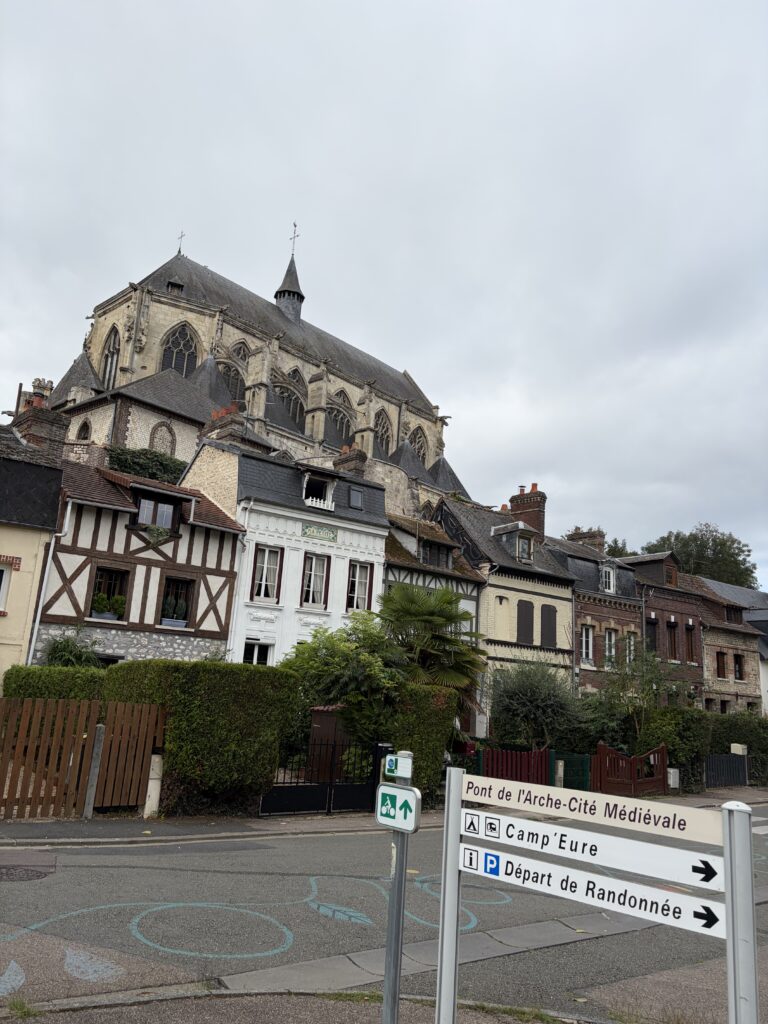 Historischer Ortskern von Pont-de-l’Arche in der Normandie mit Fachwerkhäusern und der gotischen Kirche Notre-Dame-des-Arts. Ein Wegweiser zeigt zum Campingplatz Camp’Eure und zu Wanderwegen.