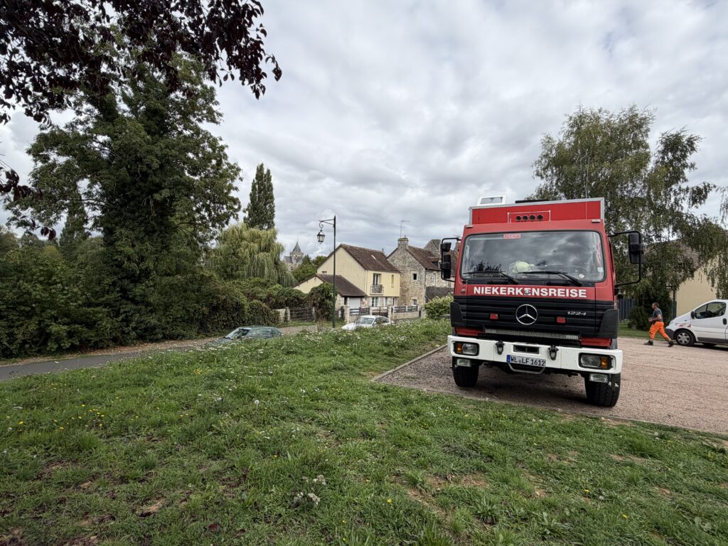 Rotes Mercedes-Benz-Expeditionsmobil von Niekerkensreise auf dem kostenlosen Wohnmobilstellplatz in Écouché-les-Vallées in der Normandie, Frankreich, umgeben von Grünflächen und historischen Häusern.