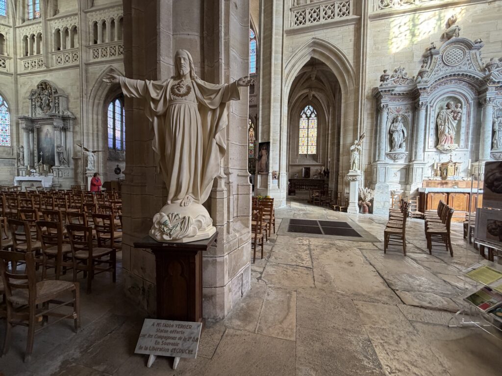 Innenraum der Kirche Saint-Martin in Écouché-les-Vallées in der Normandie mit großer Jesusstatue, gotischen Steinbögen, bunten Kirchenfenstern und kunstvoll gestalteten Seitenaltären.