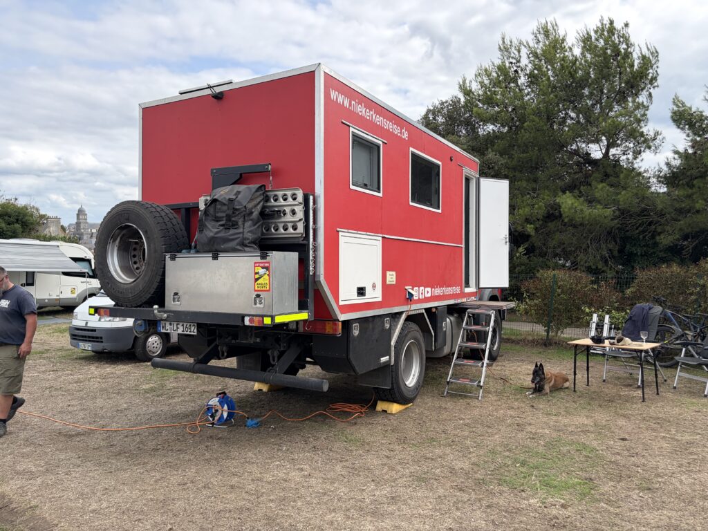 Roter Expeditions-Lkw von Niekerkensreise auf dem Campingplatz Camping de la Cité d’Alet in Saint-Malo, Bretagne. Wohnmobilstellplatz mit Stromanschluss und Hund Elvis neben dem Fahrzeug.