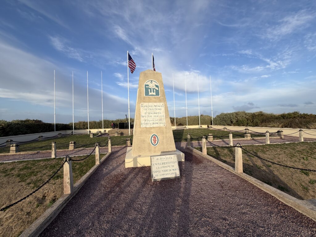 Gedenkstein der 1st Engineer Special Brigade am Utah Beach in der Normandie. Das Denkmal erinnert an die gefallenen amerikanischen Pioniere während der Landung am D-Day am 6. Juni 1944. Im Hintergrund wehen die Flaggen der USA und Frankreichs.