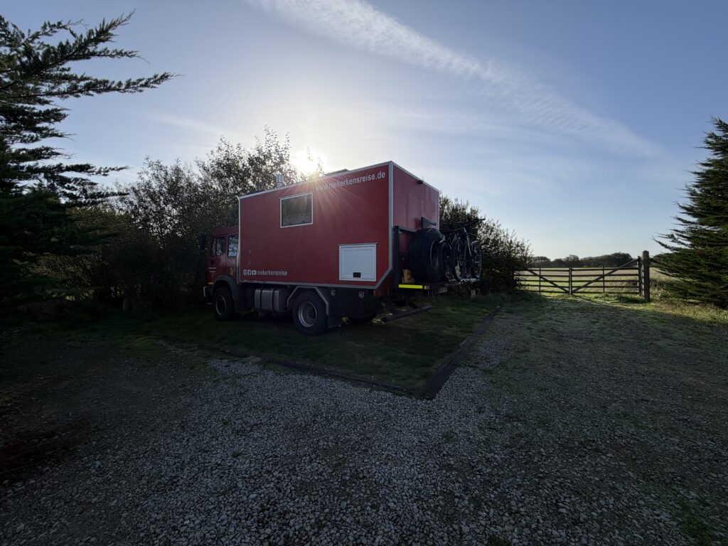 Roter Mercedes-Benz Expeditions-LKW von Niekerkensreise auf dem kostenlosen Wohnmobilstellplatz am Utah Beach in der Normandie. Das Fahrzeug steht auf einer Wiesenfläche mit Schotterzufahrt, umgeben von Bäumen und Sträuchern.