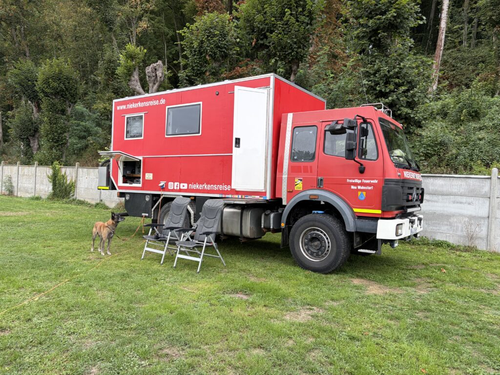 Roter Mercedes-Benz Feuerwehr-LKW mit selbst gebauter Wohnkabine von Niekerkensreise auf einer grünen Stellplatzwiese des Camping Municipal d’Étretat in der Normandie. Neben dem Expeditionsmobil stehen zwei Campingstühle und Hund Elvis.