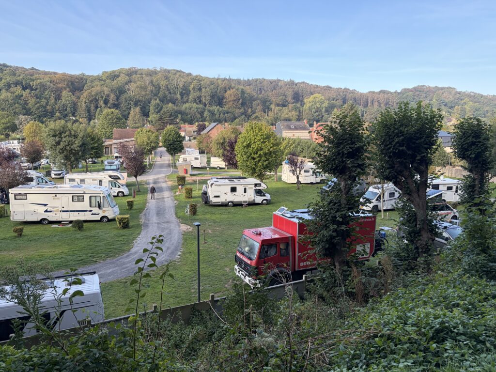 Blick über den Camping Municipal d’Étretat in der Normandie mit grünen Stellplatzwiesen, zahlreichen Wohnmobilen und dem roten Feuerwehr-LKW von Niekerkensreise. Im Hintergrund erstrecken sich bewaldete Hügel.