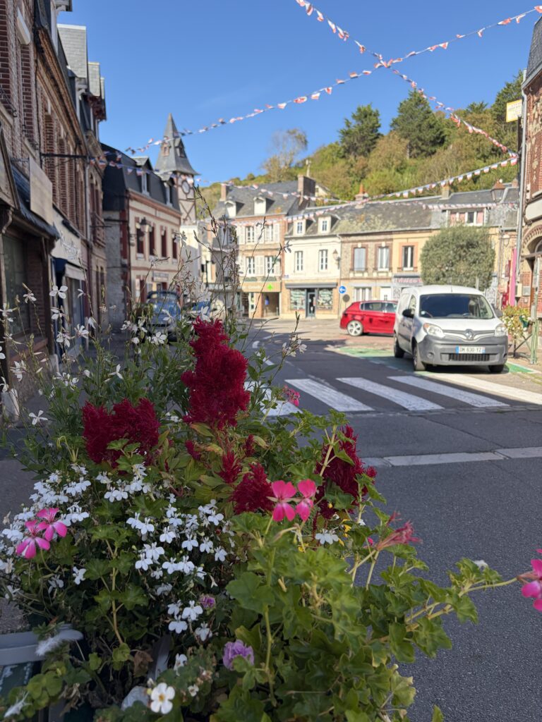 Blumengeschmückte Straße im Ortszentrum von Yport in der Normandie mit historischen Backsteinhäusern, einem markanten Uhrenturm und bunten Wimpelketten.