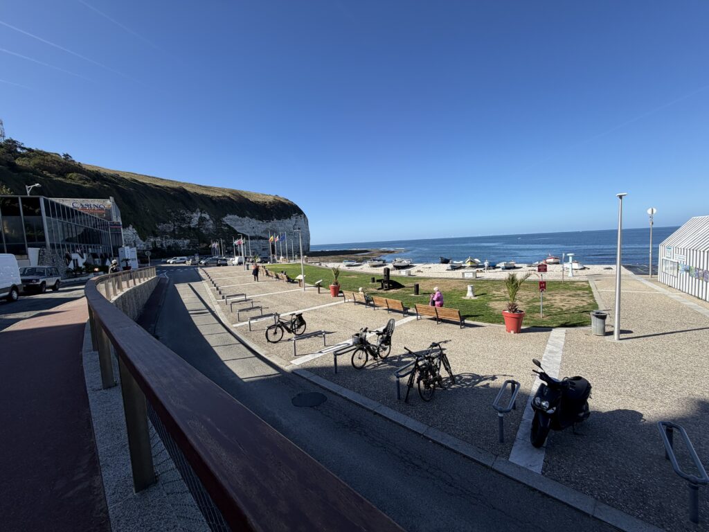 Strandpromenade von Yport in der Normandie mit Sitzbänken, Fahrrädern, Fischerbooten am Kieselstrand und den imposanten weißen Kreidefelsen der Alabasterküste bei strahlend blauem Himmel.
